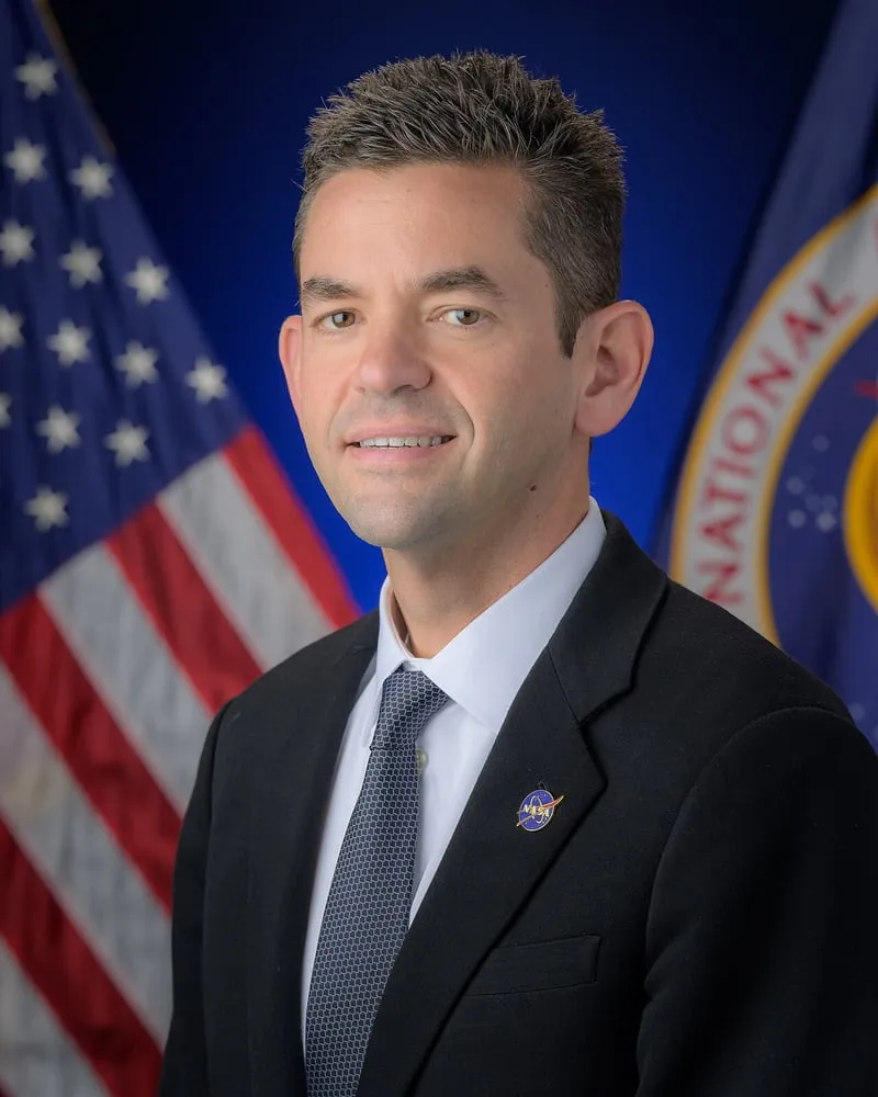 Official portrait of NASA Administrator Jared Isaacman in a dark suit, in front of the U.S. flag and the NASA flag.