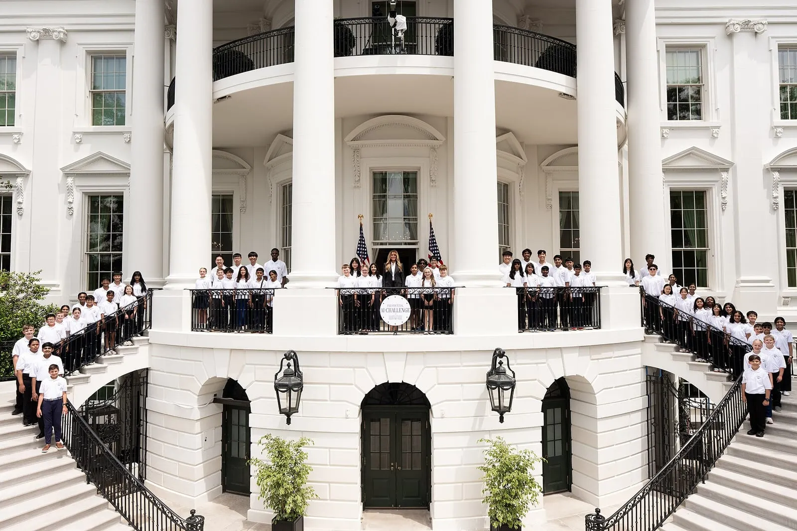 First Lady Melania Trump stands with dozens of students in white polo shirts on the White House Blue Room Balcony and its staircases, behind a 'Presidential AI Challenge' sign.